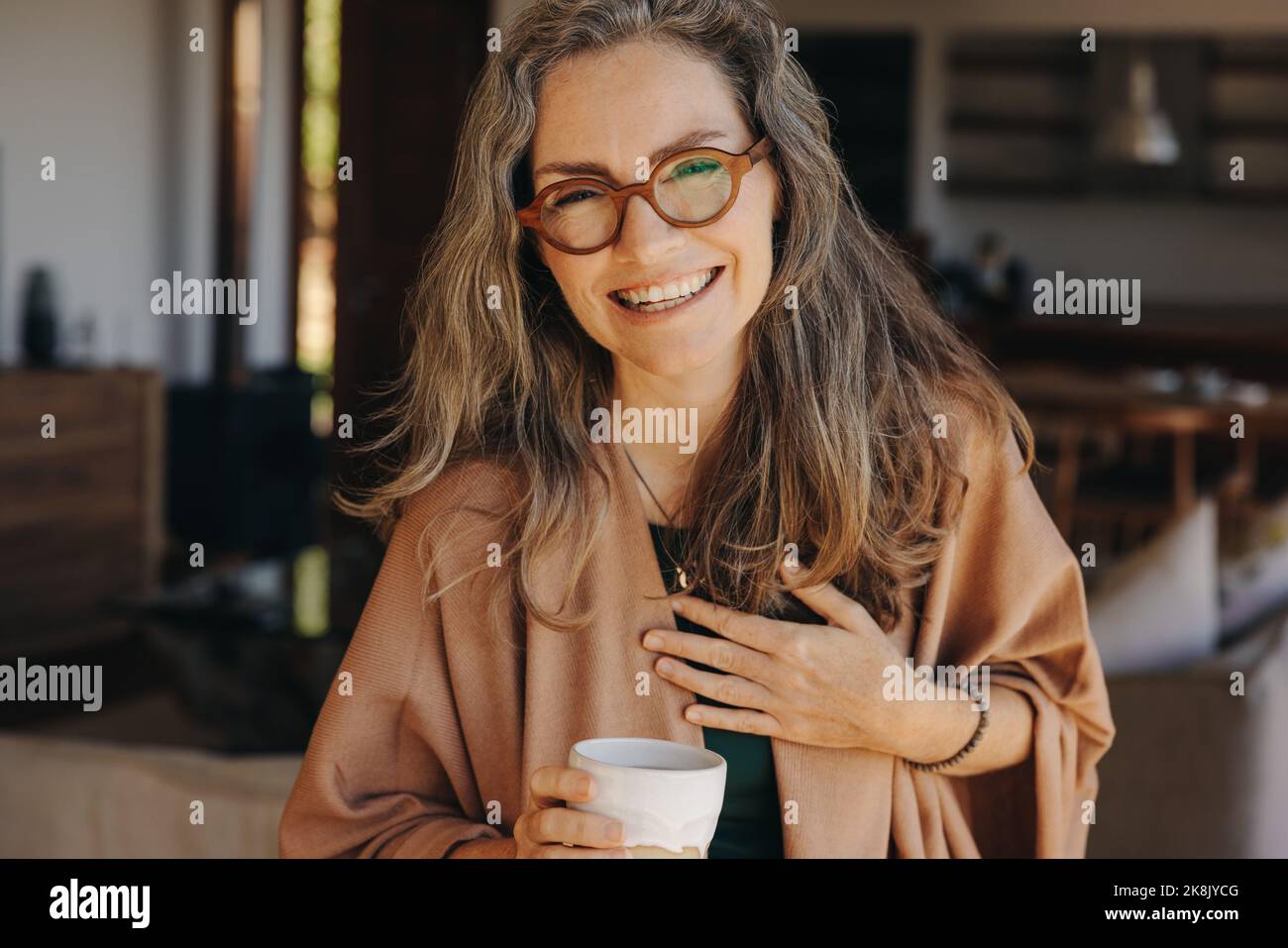 Une femme âgée gaie souriant à l'appareil photo tout en se tenant debout avec une tasse de thé dans la main. Femme mûre profitant d'une retraite heureuse à la maison. Banque D'Images