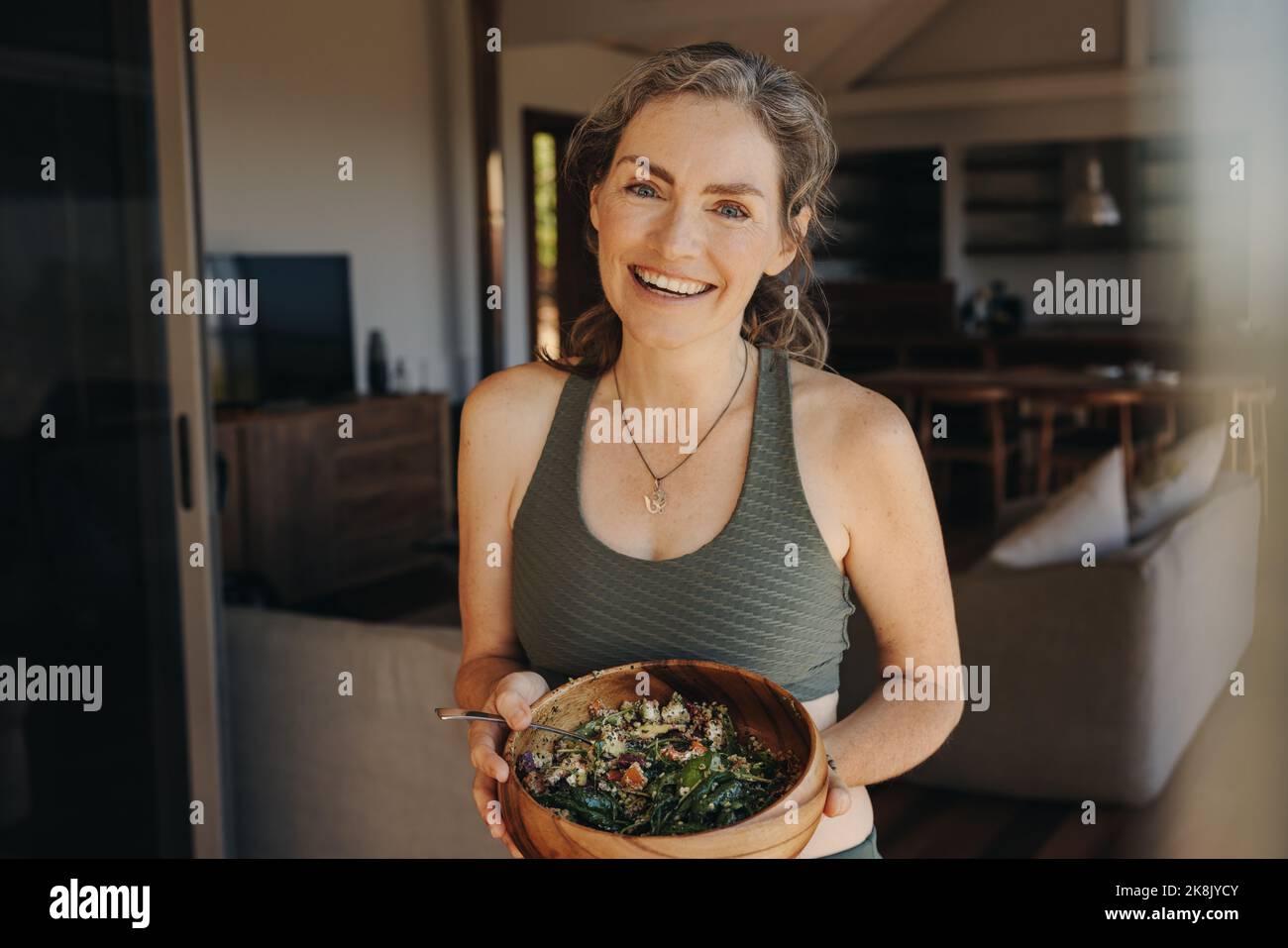 Gaie femme végétalienne souriant à l'appareil photo tout en tenant un bol de bouddha. Femme d'âge mûr prenant un petit-déjeuner à base de plantes après avoir fait de l'activité à la maison. Heureux Banque D'Images