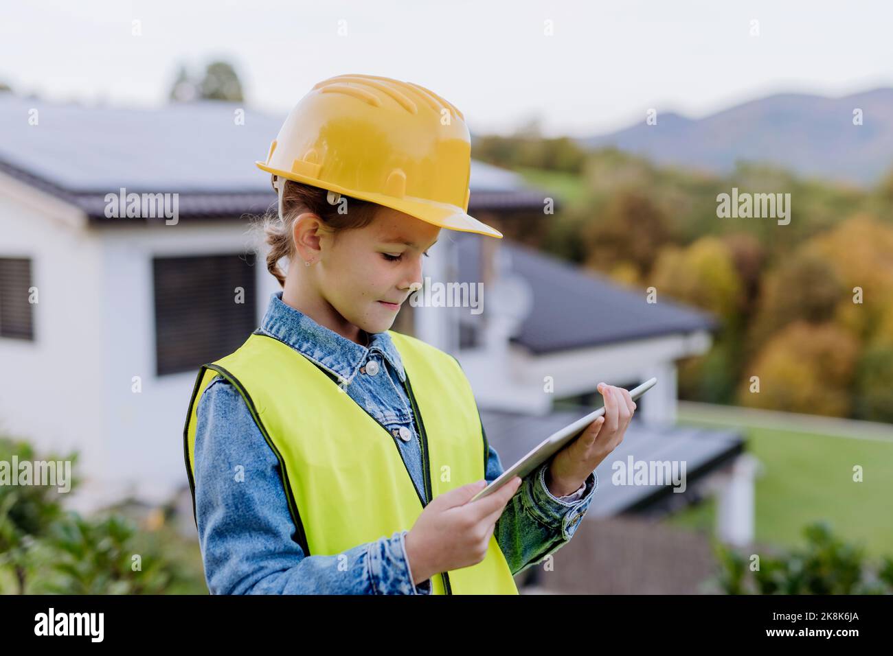 Petite fille avec casque de protection et gilet réfléchissant debout devant la maison de famille avec système solaire sur le toit et tenant la tablette numérique Banque D'Images