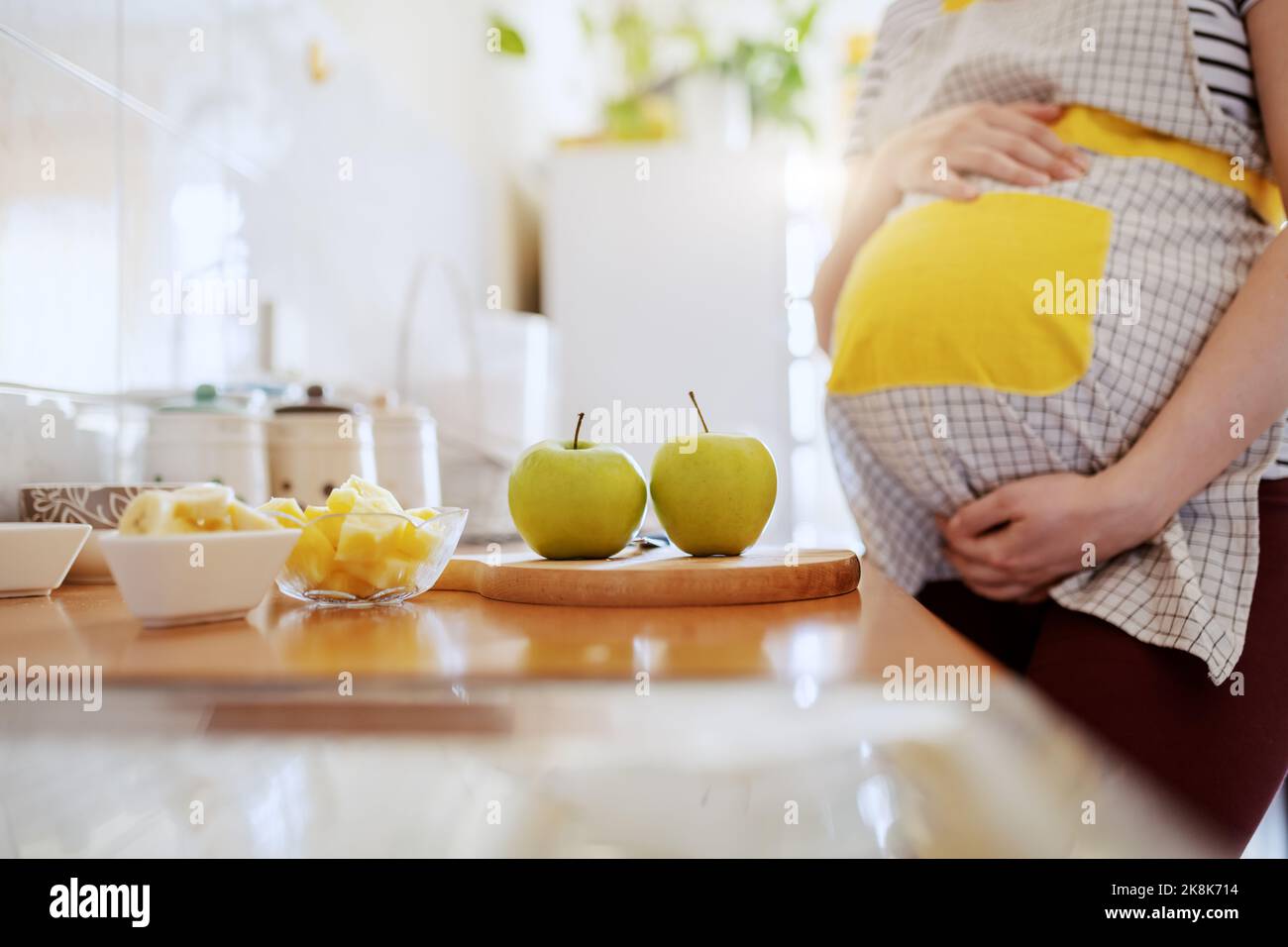Femme enceinte de race blanche en tablier debout dans la cuisine à côté du comptoir et tenant le ventre. Sur le comptoir, les pommes et les plats aux fruits. Banque D'Images