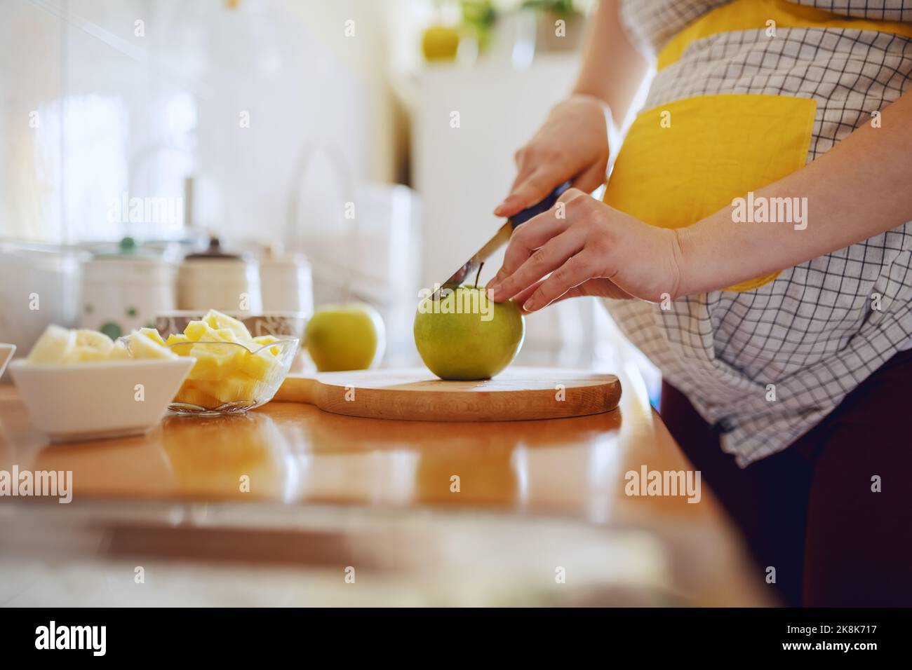 Gros plan de la femme enceinte de race blanche en tablier coupant la pomme verte dans la cuisine. Banque D'Images