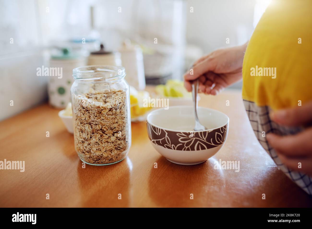 Gros plan de la femme enceinte dans un tablier debout dans la cuisine, en mettant des flocons d'avoine dans un ustensile et en préparant le petit déjeuner. Banque D'Images