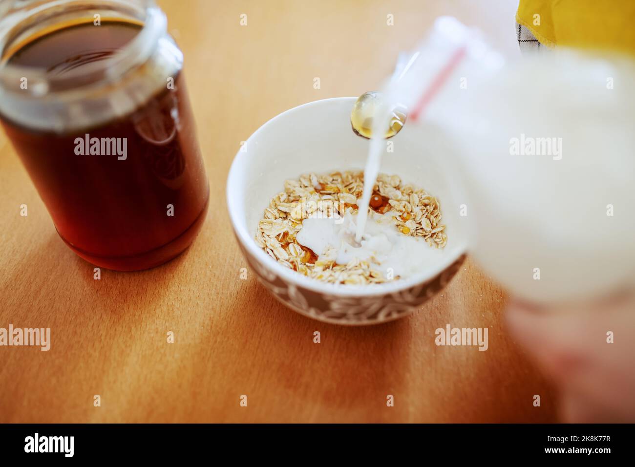 Gros plan de la femme versant du lait dans un bol avec de l'avoine. Sur le pot de table avec du miel. Banque D'Images