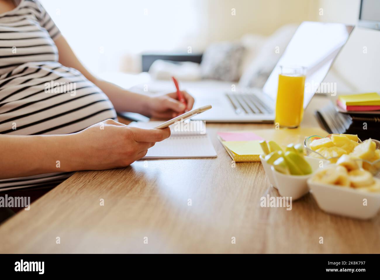 Femme enceinte assise à une table, écrivant dans un ordinateur portable et tenant un smartphone. Petit déjeuner sain et ordinateur portable à la réception. Concept de femme enceinte de travail. Banque D'Images