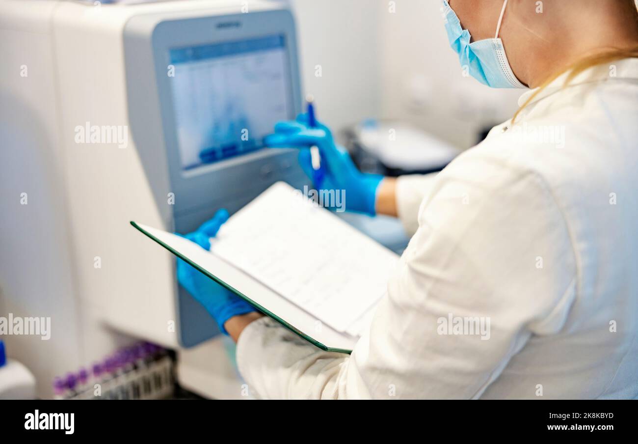 Microbiologie et biochimie. Une infirmière se tenant à côté d'une machine pour l'analyse de sang et tenant un ordre du jour avec les résultats. Elle compare les résultats si t Banque D'Images