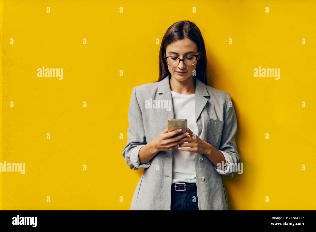Une jeune femme d'affaires se tient contre le mur jaune et utilise son téléphone pour envoyer des messages importants à son patron. WiFi, 5g, technologie Banque D'Images