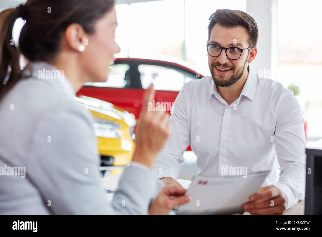 Sourire le client assis à la table avec le vendeur de voiture, tenant l'offre et écoutant le vendeur de voiture au sujet des commodités. Banque D'Images
