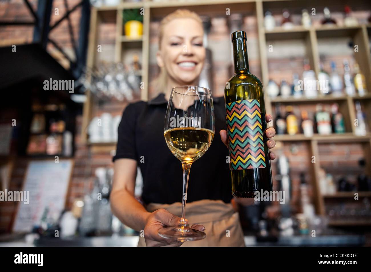 Concentrez-vous sur le vin au bar. Vue de face d'une femme connaisseur de vin dans un uniforme moderne qui travaille derrière le bar. Elle tient une bouteille de vin dans une main un Banque D'Images