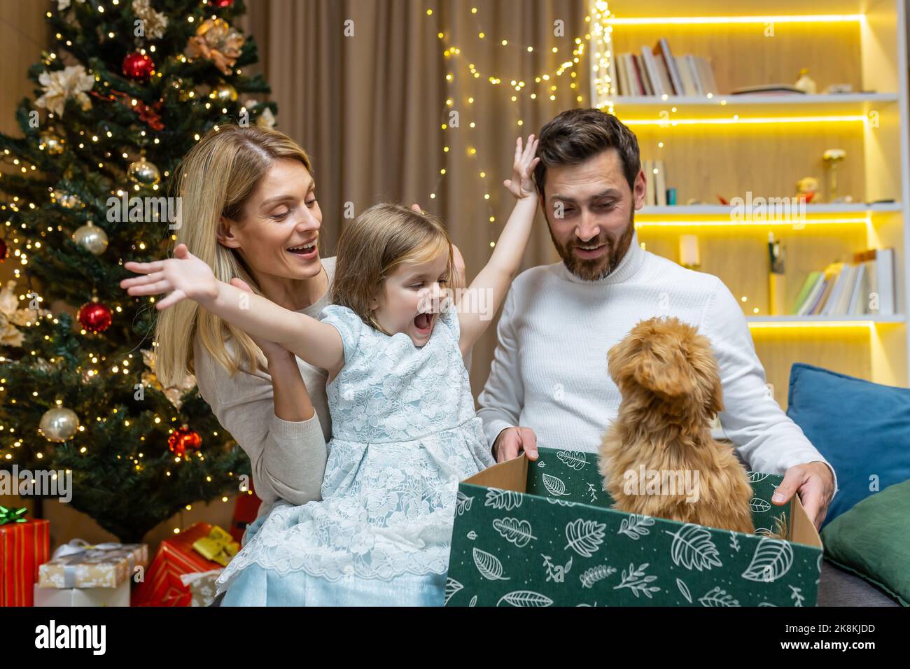 Bonne famille du nouvel an pour Noël, mari et femme avec fille dans le salon près de l'arbre de Noël, partageant des cadeaux, donnant un petit chien de maltipoo d'animal de compagnie à la fille. Banque D'Images