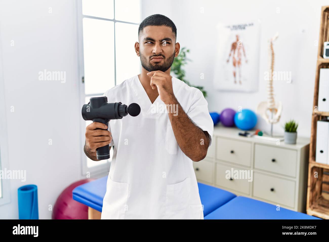 Jeune kinésithérapeute indien tenant un pistolet de massage thérapeutique au centre de bien-être pensant inquiet à une question, inquiet et nerveux avec la main sur le menton Banque D'Images