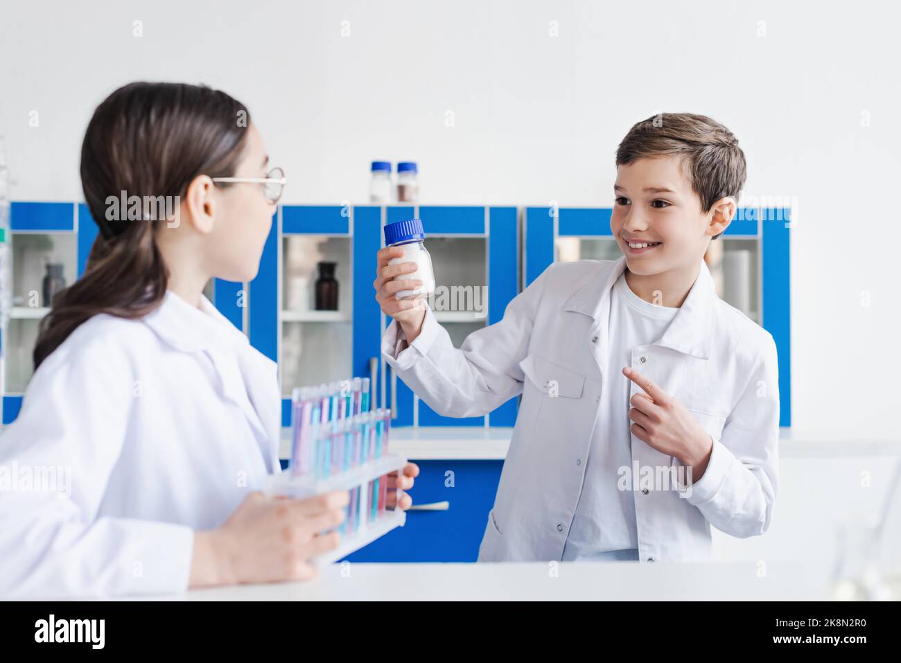 joyeux garçon pointant vers le pot avec substance blanche près de la fille floue avec des tubes à essai, image de stock Banque D'Images