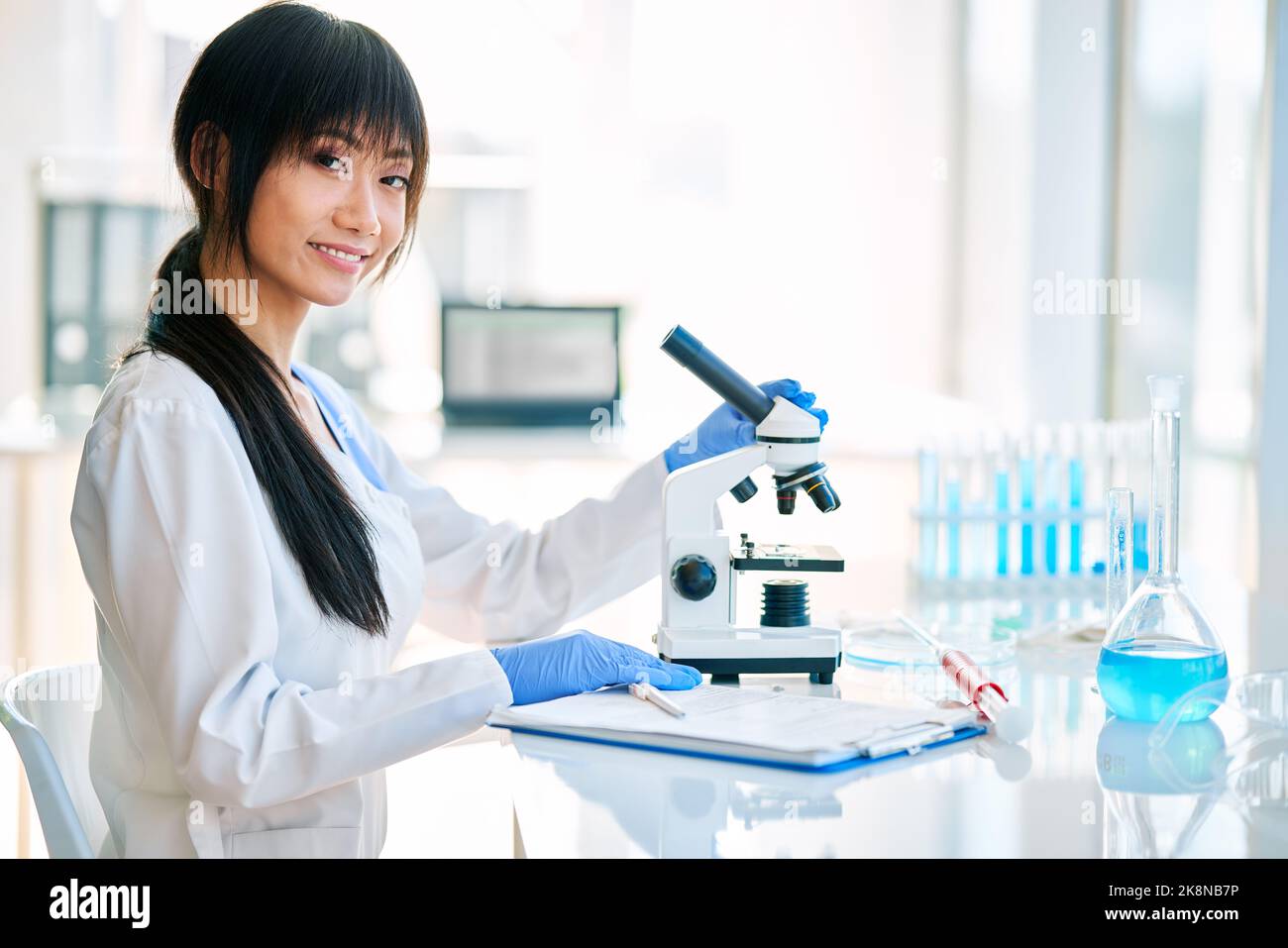 Une scientifique asiatique souriante se pose dans un laboratoire de recherche médicale moderne à côté d'un microscope. Banque D'Images