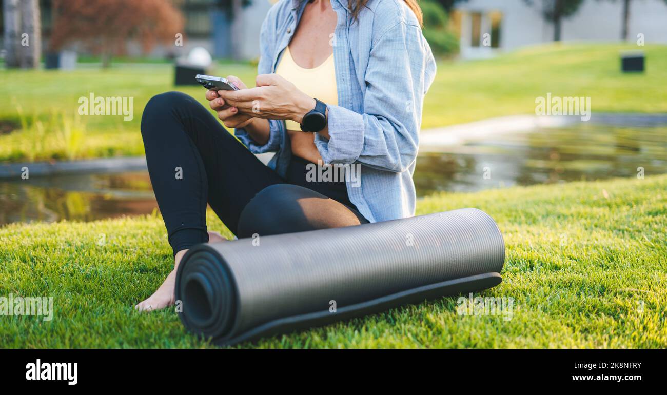 Vue rognée d'une femme regardant une vidéo sur un smartphone assis sur une surface herbeuse, en appréciant l'heure du matin. Femme utilisant son smartphone pour envoyer un SMS Banque D'Images