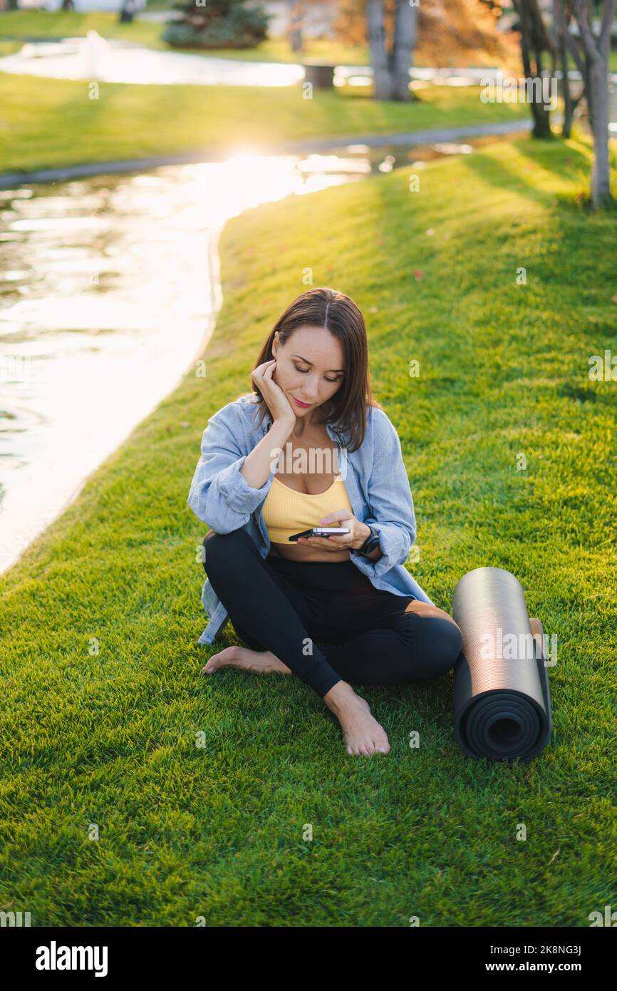 Pleine longueur d'une femme sportive tenant un smartphone assis sur la pelouse verte près d'une rivière. Un mode de vie sain et actif. Entraînement physique. Fitness Banque D'Images