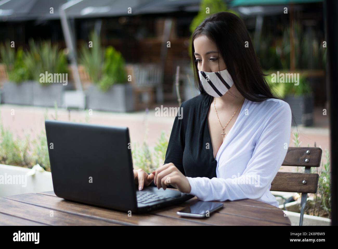 Une femme portant un masque, travaillant sur son ordinateur portable avec une expression sérieuse dans un café Banque D'Images