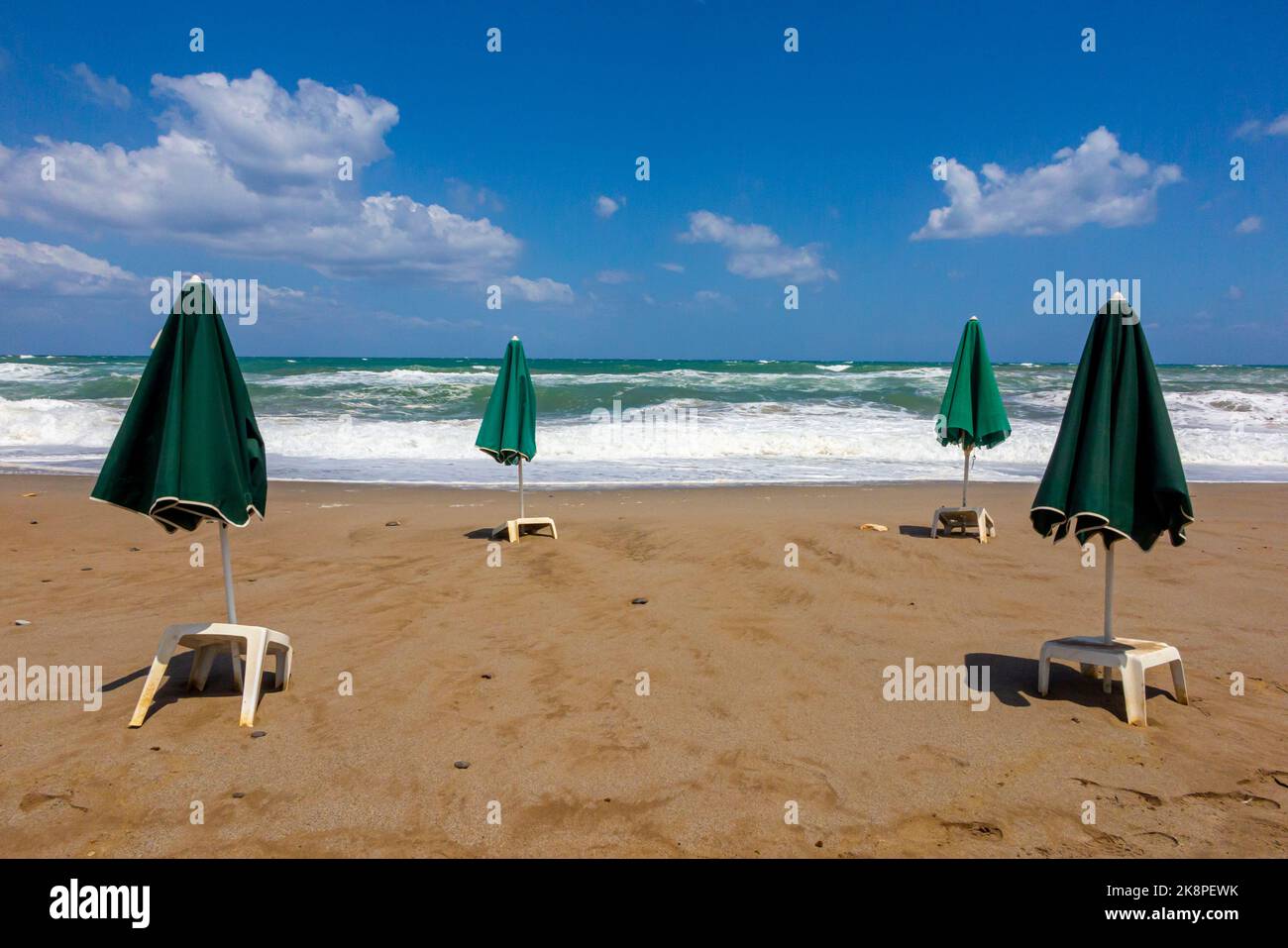 Parasols verts et tables en plastique blanc sur une plage de sable déserte avec des vagues s'écrasant sur le sable en arrière-plan et ciel bleu avec des nuages au-dessus. Banque D'Images