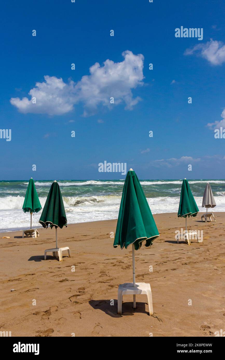Parasols verts et tables en plastique blanc sur une plage de sable déserte avec des vagues s'écrasant sur le sable en arrière-plan et ciel bleu avec des nuages au-dessus. Banque D'Images
