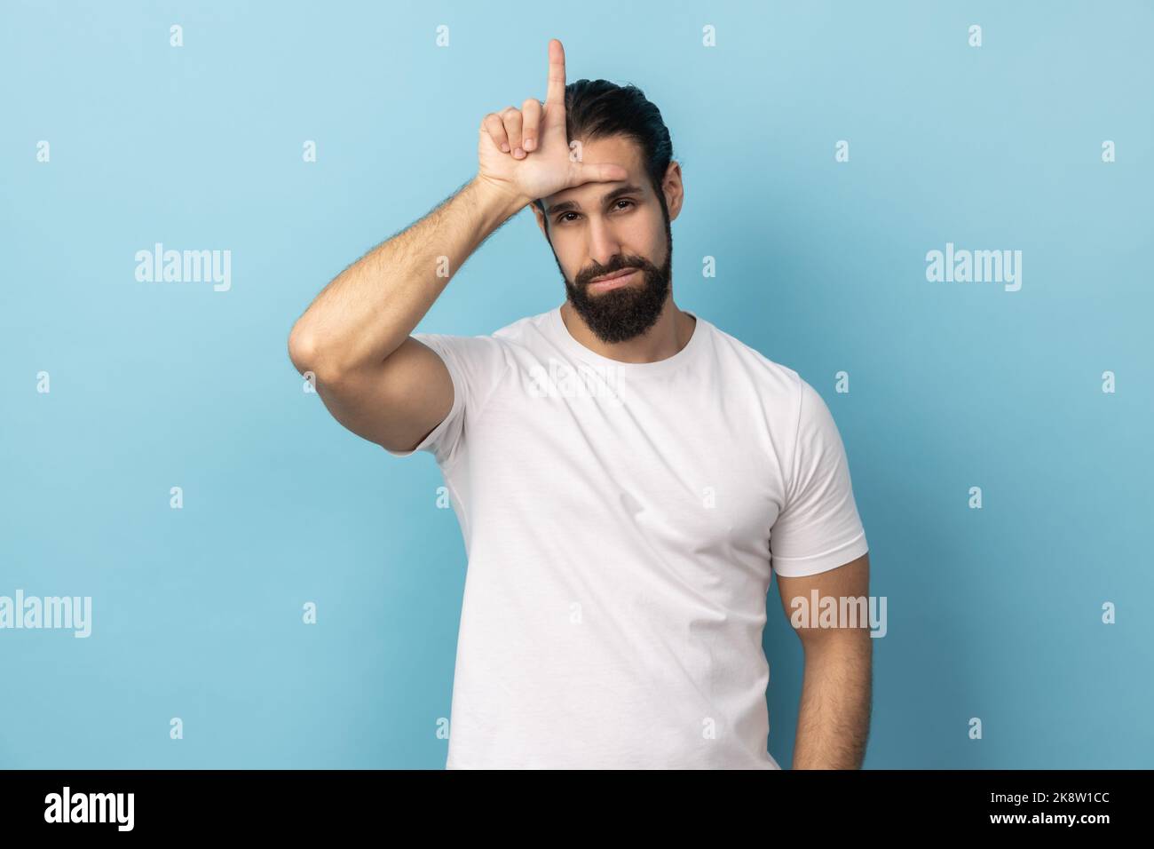 J'ai perdu mon travail. Portrait d'un homme avec une barbe portant un T-shirt blanc montrant un geste perdant, L signe du doigt sur le front, contrarié par le congédiement, jour malchanceux. Studio d'intérieur isolé sur fond bleu. Banque D'Images