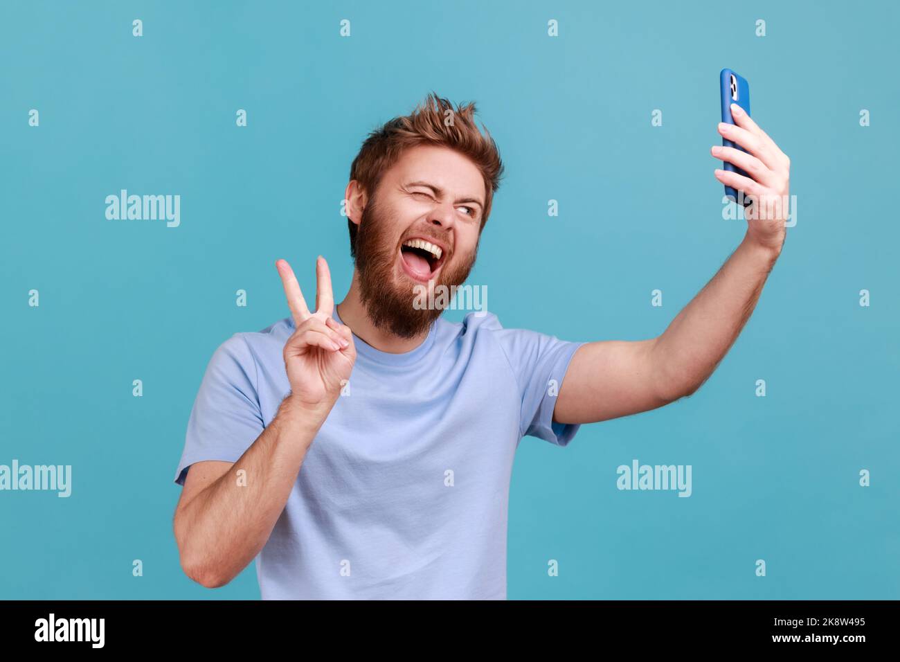 Portrait d'un homme réussi prenant le selfie ou parlant sur appel vidéo et montrant la victoire, geste de paix, agréable communication en ligne via l'application de téléphone mobile. Studio d'intérieur isolé sur fond bleu Banque D'Images