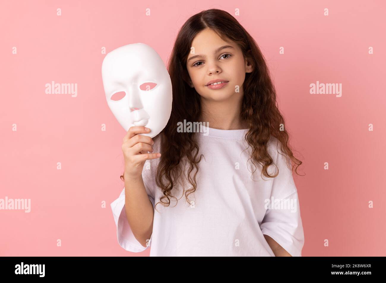 Portrait de charmante adorable adorable petite fille à cheveux foncés portant un T-shirt blanc tenant un masque blanc dans sa main, veut changer de personnalité. Studio d'intérieur isolé sur fond rose. Banque D'Images