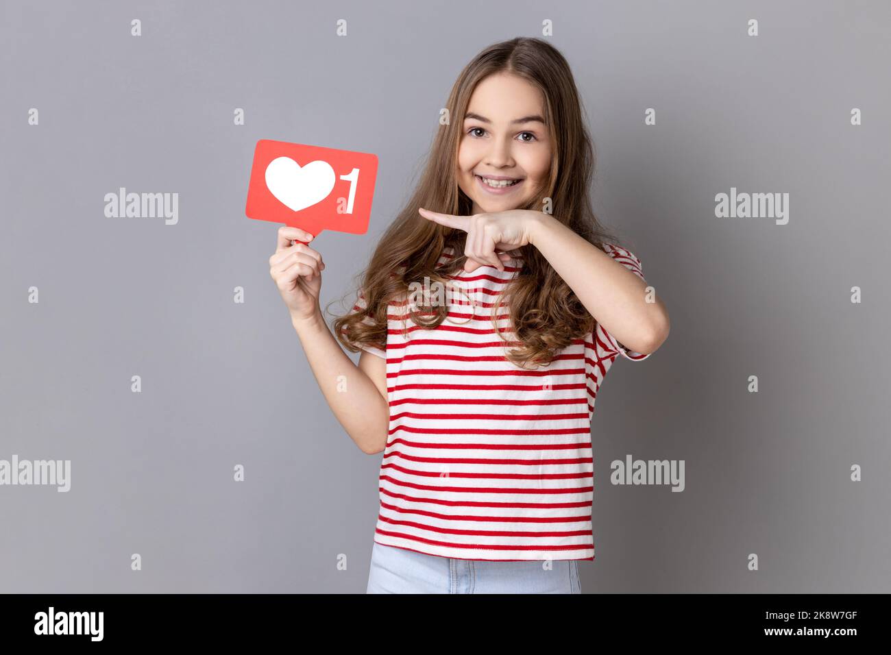 Portrait d'une adorable petite fille souriante portant un T-shirt rayé pointant vers le cœur comme une icône, recommandant de cliquer sur le bouton des médias sociaux. Prise de vue en studio isolée sur fond gris. Banque D'Images