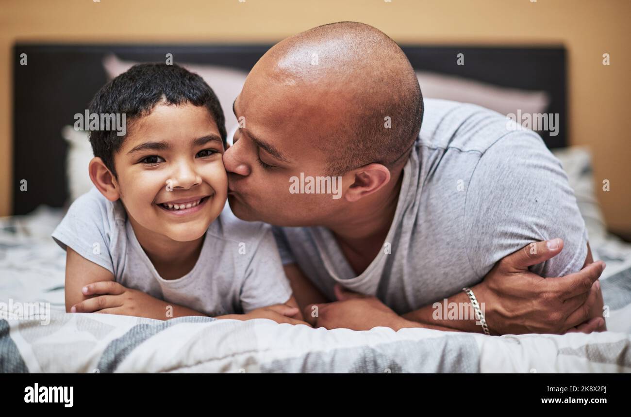 Mon grand garçon. Portrait d'un petit garçon joyeux recevant un baiser sur sa joue par son père pendant qu'ils traînent sur le lit à la maison pendant la journée. Banque D'Images