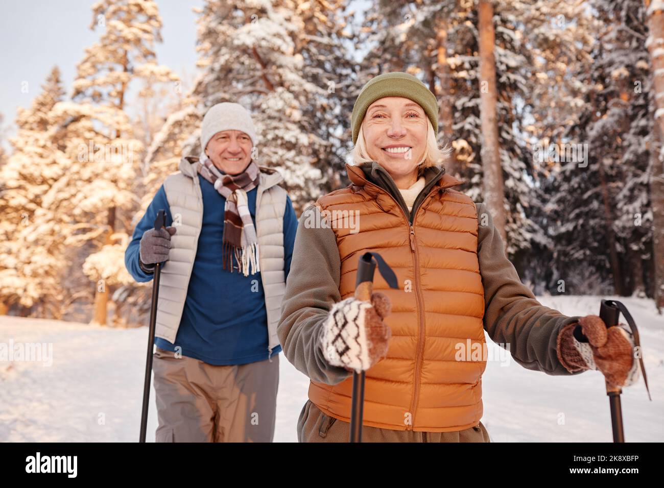 Portrait à la taille du couple senior actif marchant avec des bâtons dans la forêt d'hiver et souriant à la caméra Banque D'Images