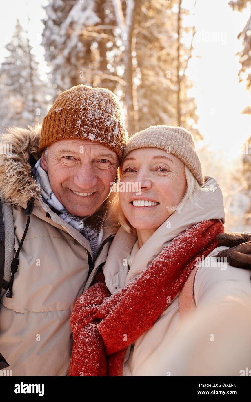 Plan d'ensemble vertical d'un couple senior heureux prenant des photos de selfie tout en appréciant la randonnée dans la forêt d'hiver Banque D'Images