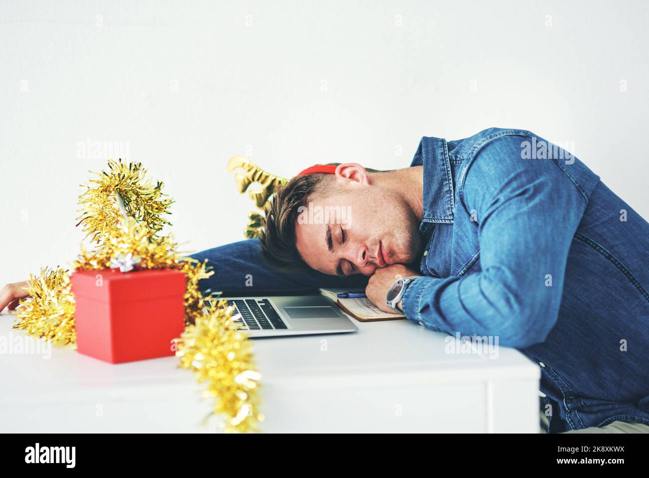 Travailler pendant les fêtes est un vrai tracas : un beau jeune homme d'affaires qui dormait sur son bureau pendant qu'il travaillait dans son bureau la veille de Noël. Banque D'Images