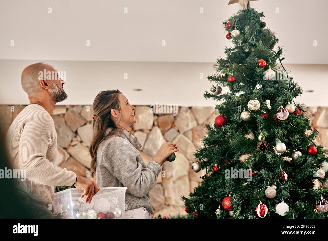Il n'y a presque pas de place pour mettre cela. Un couple joyeux d'âge moyen plaçant des décorations sur un arbre de Noël à l'intérieur à la maison pendant la journée. Banque D'Images