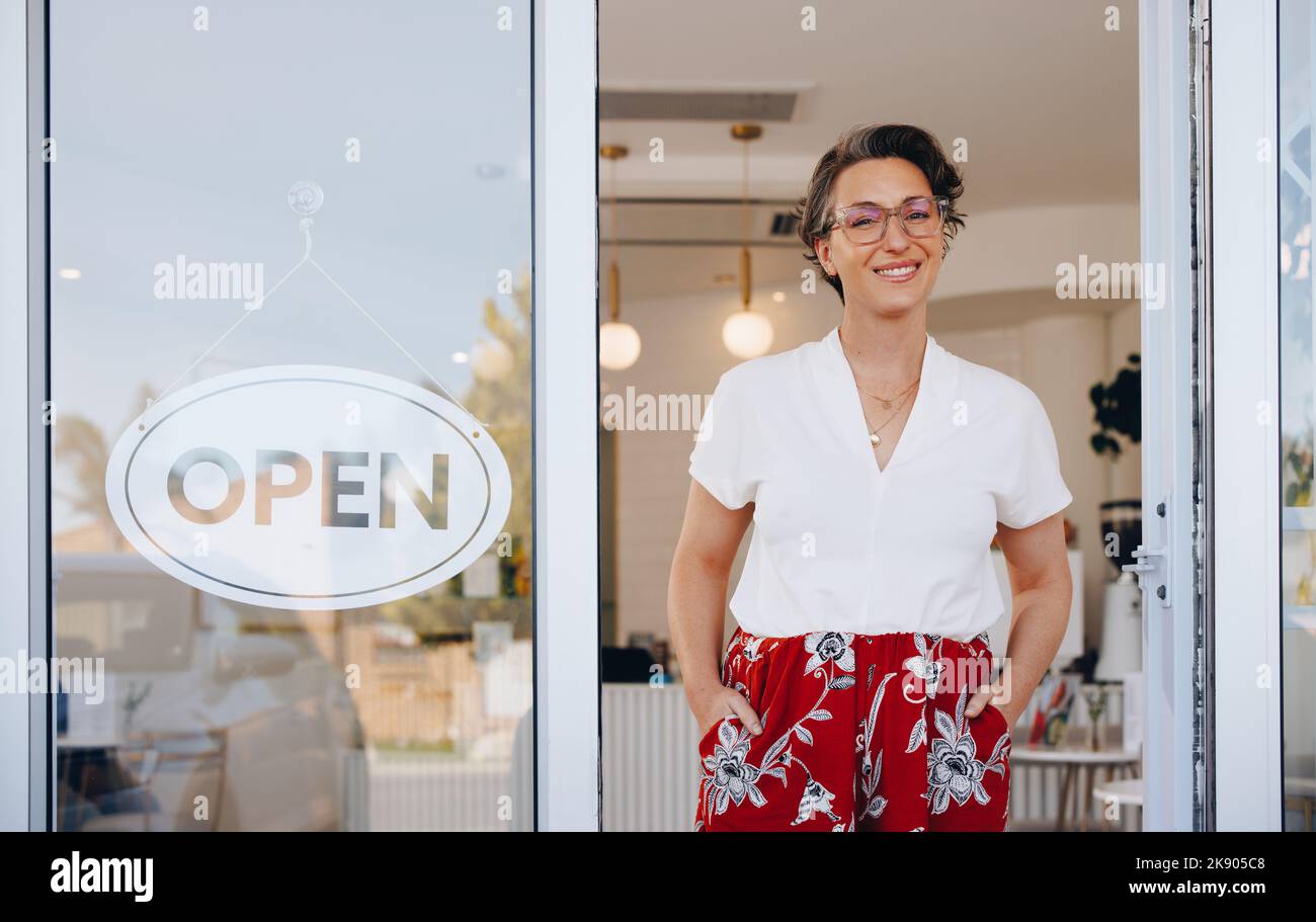 Femme mûre souriant à la caméra tout en se tenant à côté d'un panneau ouvert à la porte de son restaurant. Un propriétaire de petite entreprise heureux accueille le client Banque D'Images