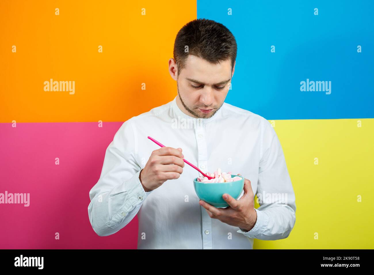 Un homme sur fond jaune, bleu, orange, rose dans une chemise blanche avec un plateau turquoise de guimauves et une cuillère rose. Le gars avec un sourire. Un im Banque D'Images