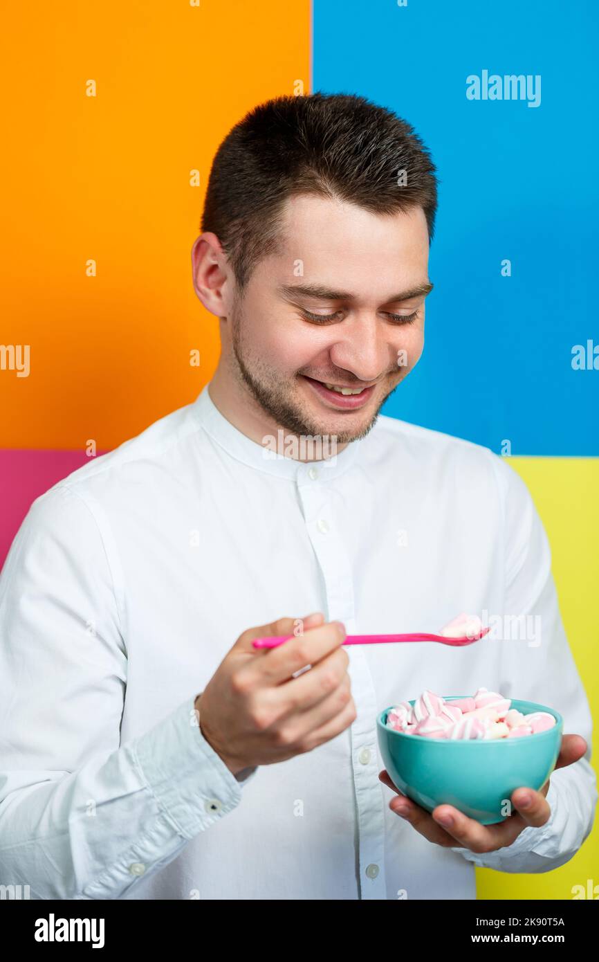 Un homme sur fond jaune, bleu, orange, rose dans une chemise blanche avec un plateau turquoise de guimauves et une cuillère rose. Le gars avec un sourire. Un im Banque D'Images