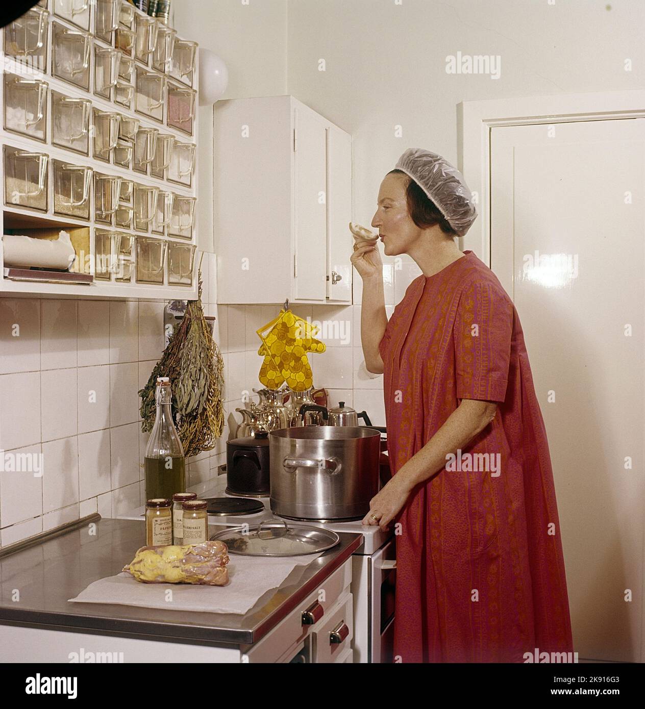 Dans la cuisine 1960s. Intérieur d'une cuisine et d'une femme de cuisine debout près du poêle dégustation de quelque chose des casseroles et des poêles. Elle a le filet à cheveux sur pour empêcher les cheveux tomber dans la nourriture. Suède 1962 réf. cv83 Banque D'Images