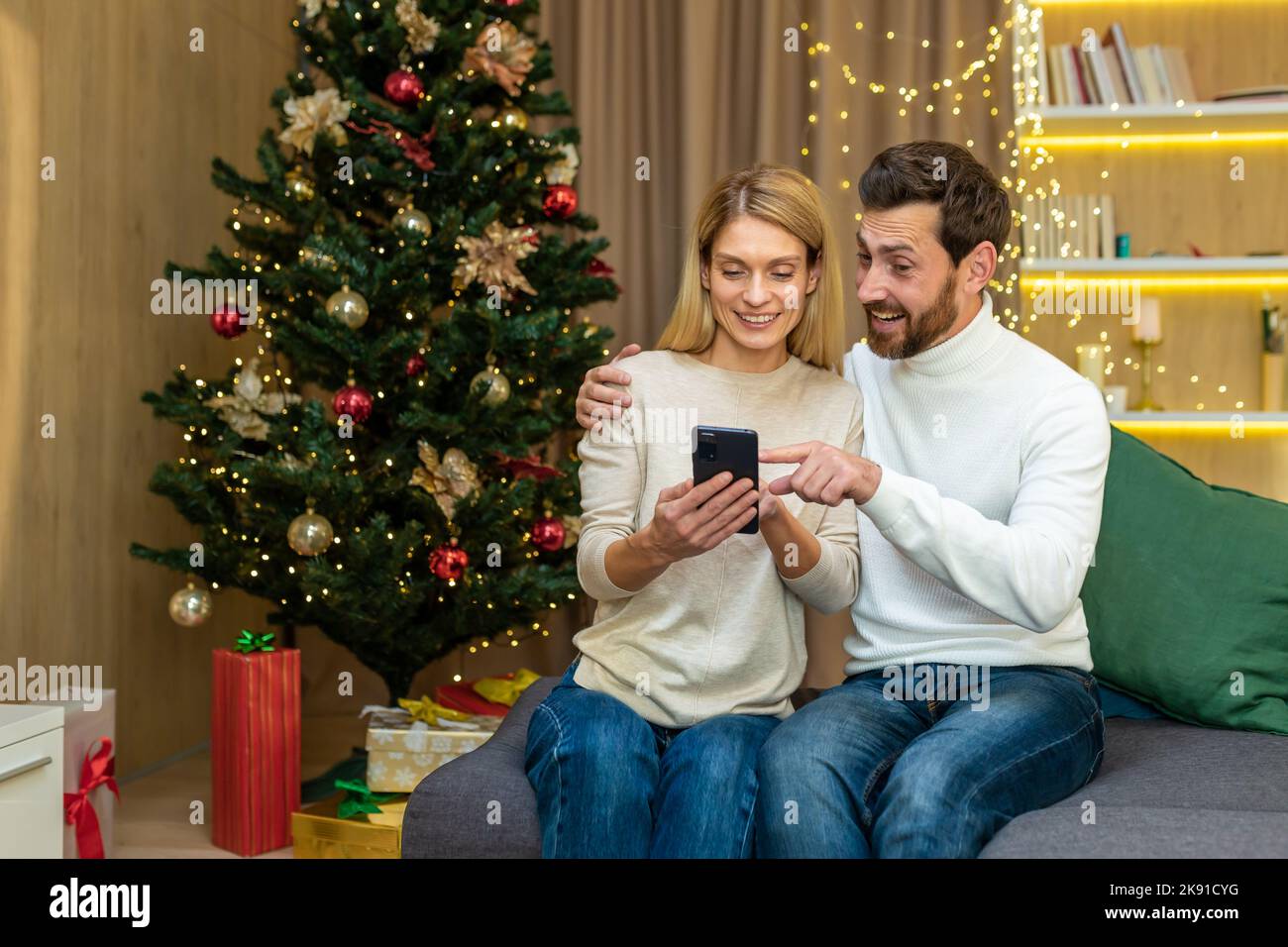 Couple de famille en amour homme et femme pour noël célébrant le nouvel an assis sur le canapé à la maison regarder la vidéo sur le téléphone et sourire en choisissant des cadeaux dans la télécommande de magasin en ligne. Banque D'Images