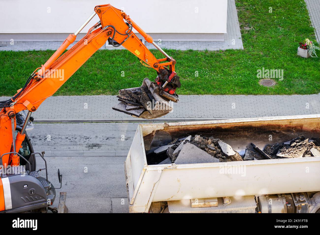 Une pelle hydraulique orange équipée d'un godet charge les anciens morceaux d'asphalte dans un camion à benne blanche Banque D'Images