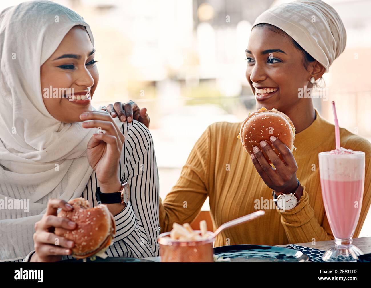 Les gourmets savent comment se rendre heureux. Deux jeunes amies affectueuses mangeant des hamburgers dans un café tout en étant vêtues de hijab. Banque D'Images