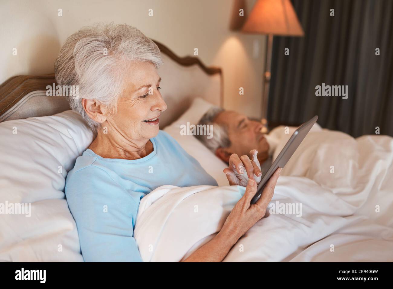 En regardant le profil en ligne de mes filles, une femme âgée assise au lit et utilisant une tablette pendant que son mari dort à côté d'elle. Banque D'Images