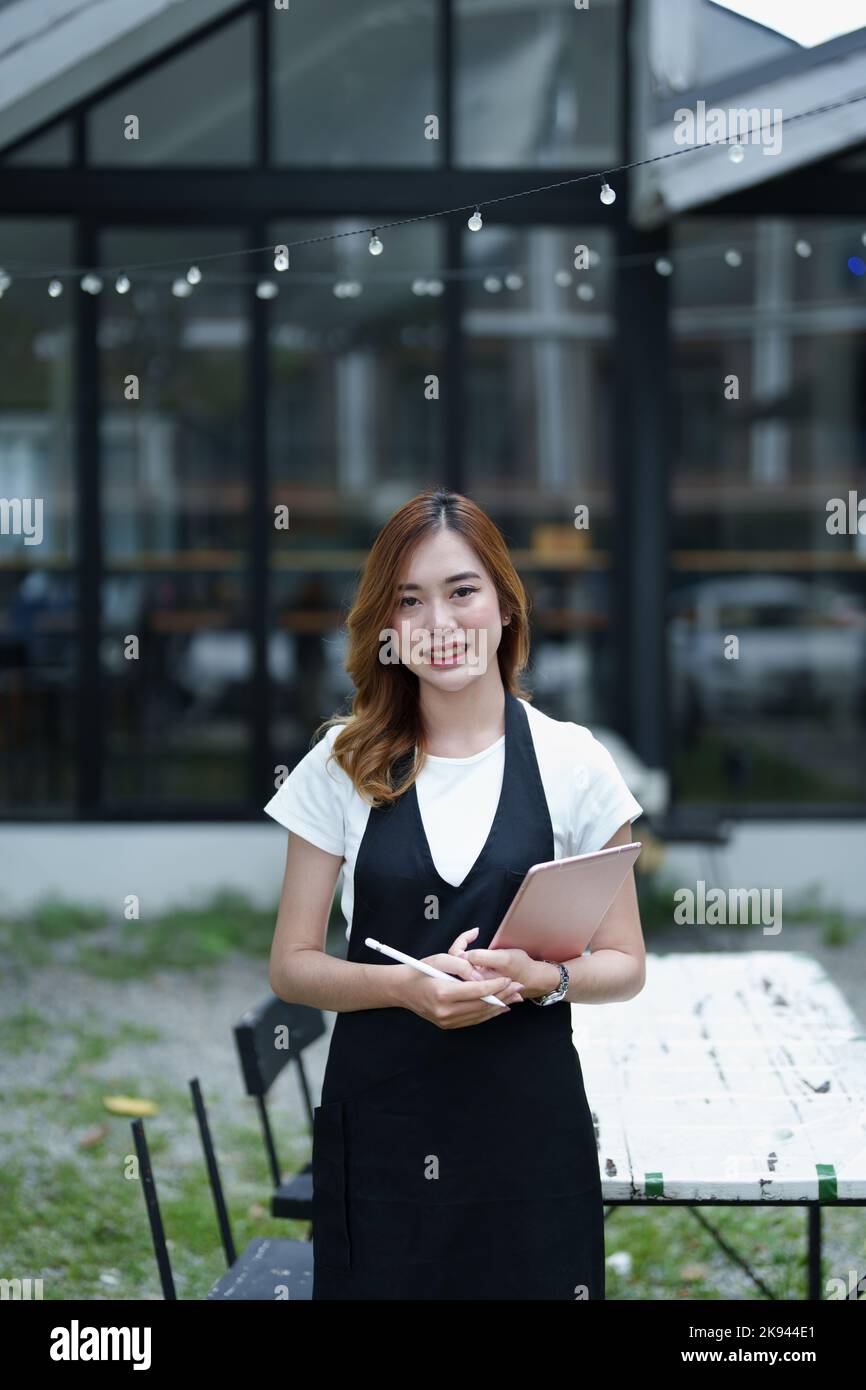 Début et ouverture d'une petite entreprise, une jeune femme asiatique montrant un visage souriant tenant une tablette dans un tablier devant un café-bar Banque D'Images