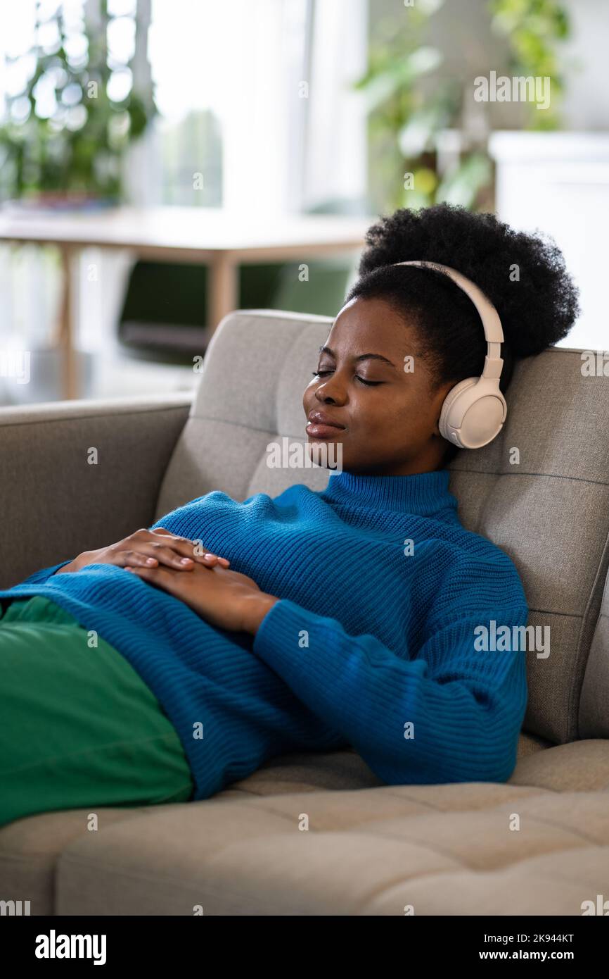 Une femme afro-américaine qui se repose dans un casque avec un petit sourire se trouve sur un canapé qui s'endorme à la musique Banque D'Images