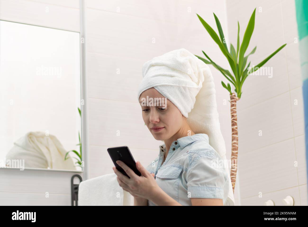 Une jeune femme avec une serviette blanche sur sa tête dans une salle de bains lumineuse communique avec un smartphone avec un ami dans les réseaux sociaux.Réflexion dans le m Banque D'Images