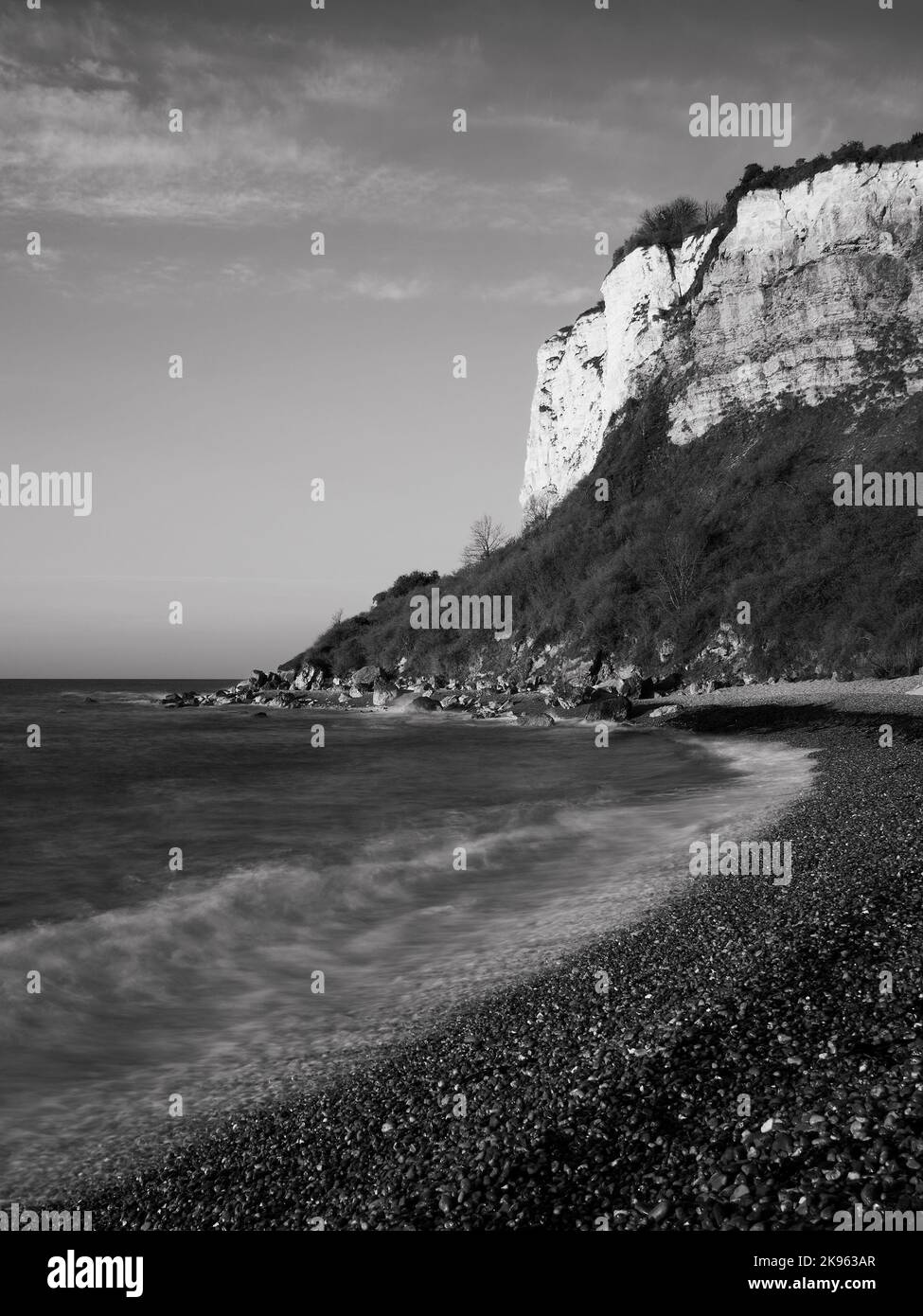 Exposition longue, convertie en noir et blanc, de vagues qui se jettent sur la plage de galets en face des falaises de craie au lever du soleil. La côte jurassique, les plus vantent Banque D'Images