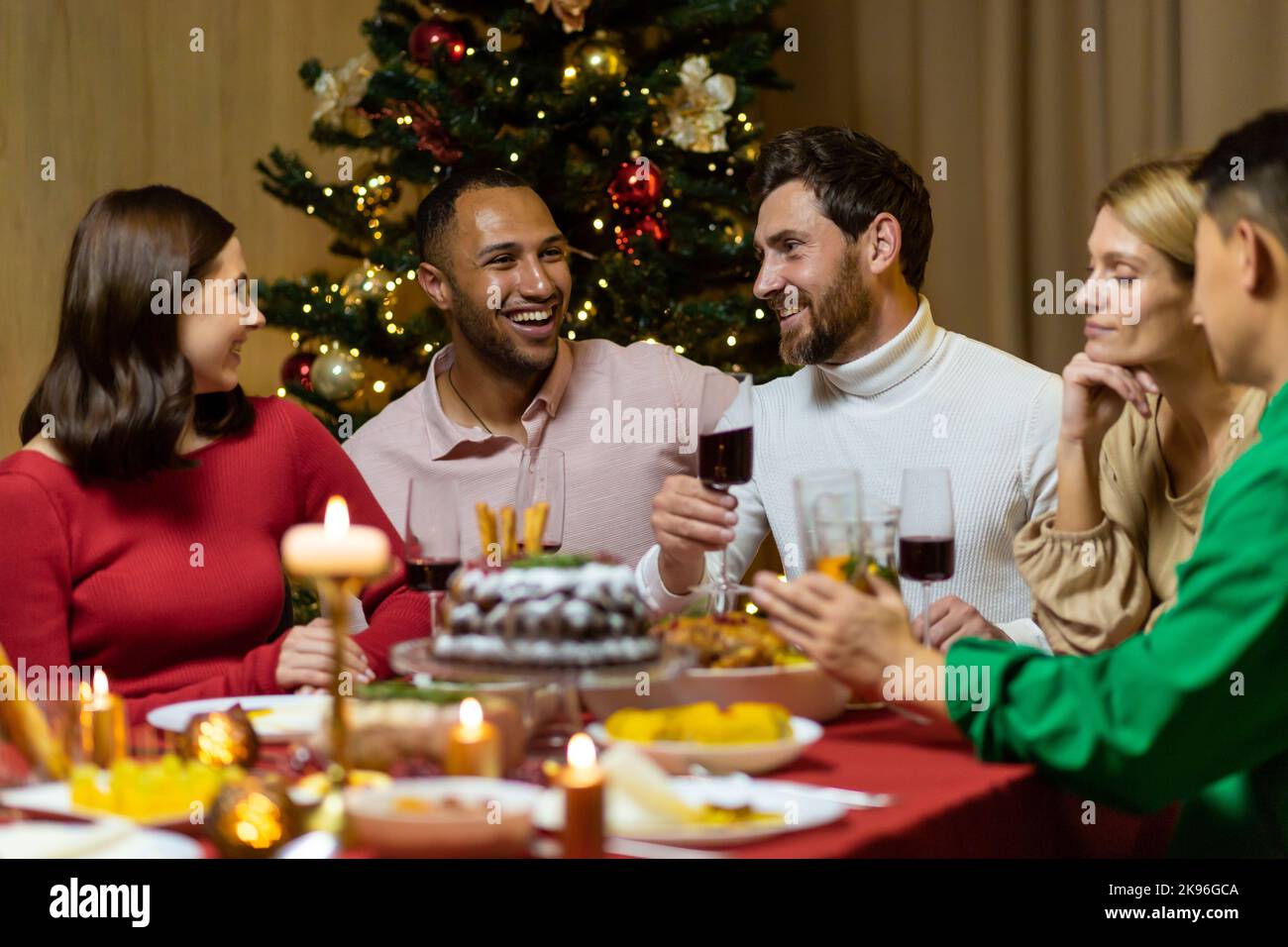 Les amis fêtent la veille de Noël ou le jour de l'an ensemble assis à la table. Dînez à la maison avec un dîner de Noël multiethnique. Joyeux et joyeux, les gens rient avec des verres à vin Banque D'Images