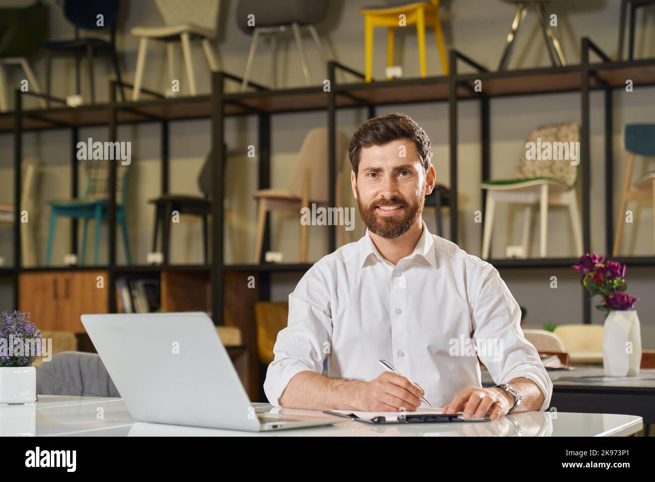 Un détaillant homme confiant travaillant sur un ordinateur portable, tout en souriant dans un magasin de meubles moderne. Portrait d'un entrepreneur heureux prenant des notes, contre un rack avec plusieurs chaises sur l'arrière-plan. Concept de lieu de travail. Banque D'Images Un détaillant homme confiant travaillant sur un ordinateur portable, tout en souriant dans un magasin de meubles moderne. Portrait d'un entrepreneur heureux prenant des notes, contre un rack avec plusieurs chaises sur l'arrière-plan. Concept de lieu de travail. Banque D'Images