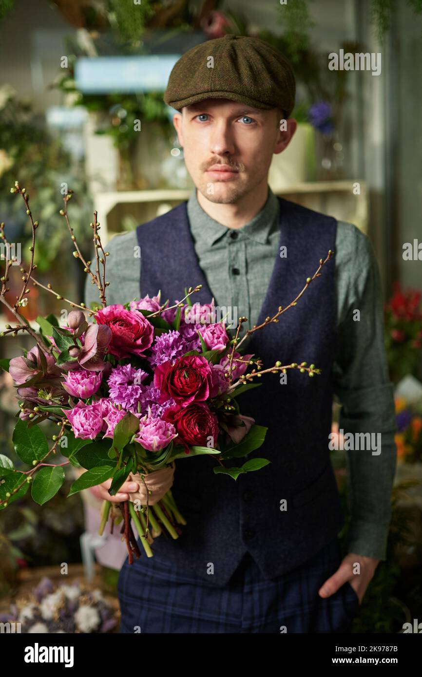 Concept de la Saint-Valentin ou de la Journée des femmes. Attrayant mâle en casquette à visière et vêtements vintage de 20s tenant beau bouquet de fleurs rouges et violettes. Félicitez avec des fleurs. Image de haute qualité Banque D'Images