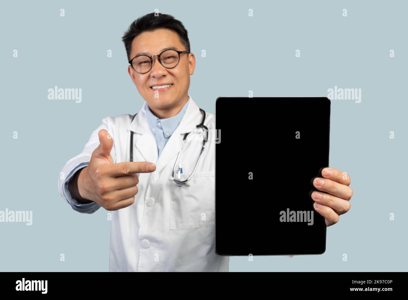 Thérapeute chinois mature satisfait masculin en manteau blanc, lunettes pointant sur un comprimé avec écran vide Banque D'Images