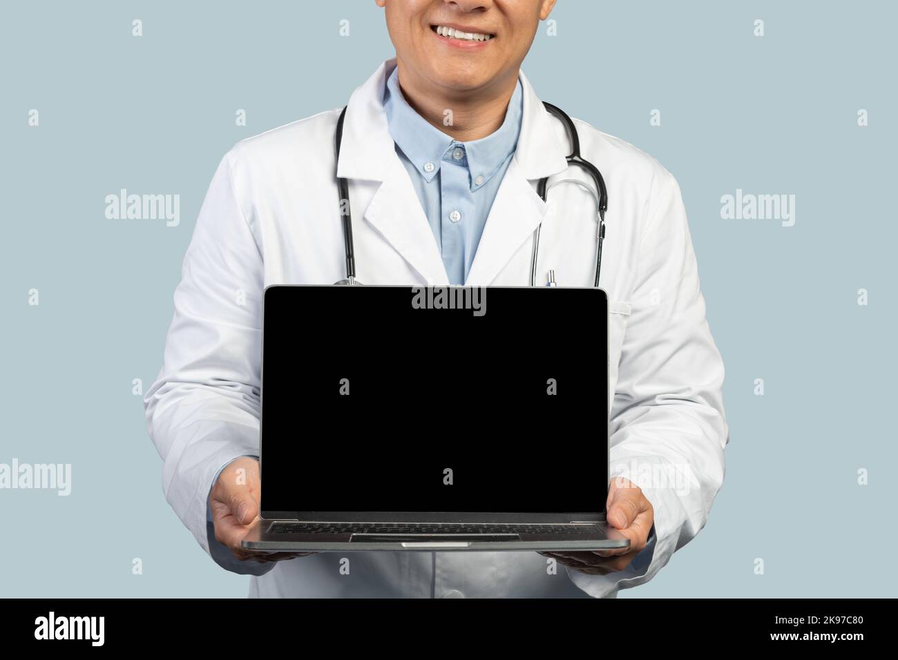 Court heureux coréen d'âge moyen médecin en manteau blanc montre ordinateur portable avec écran blanc Banque D'Images