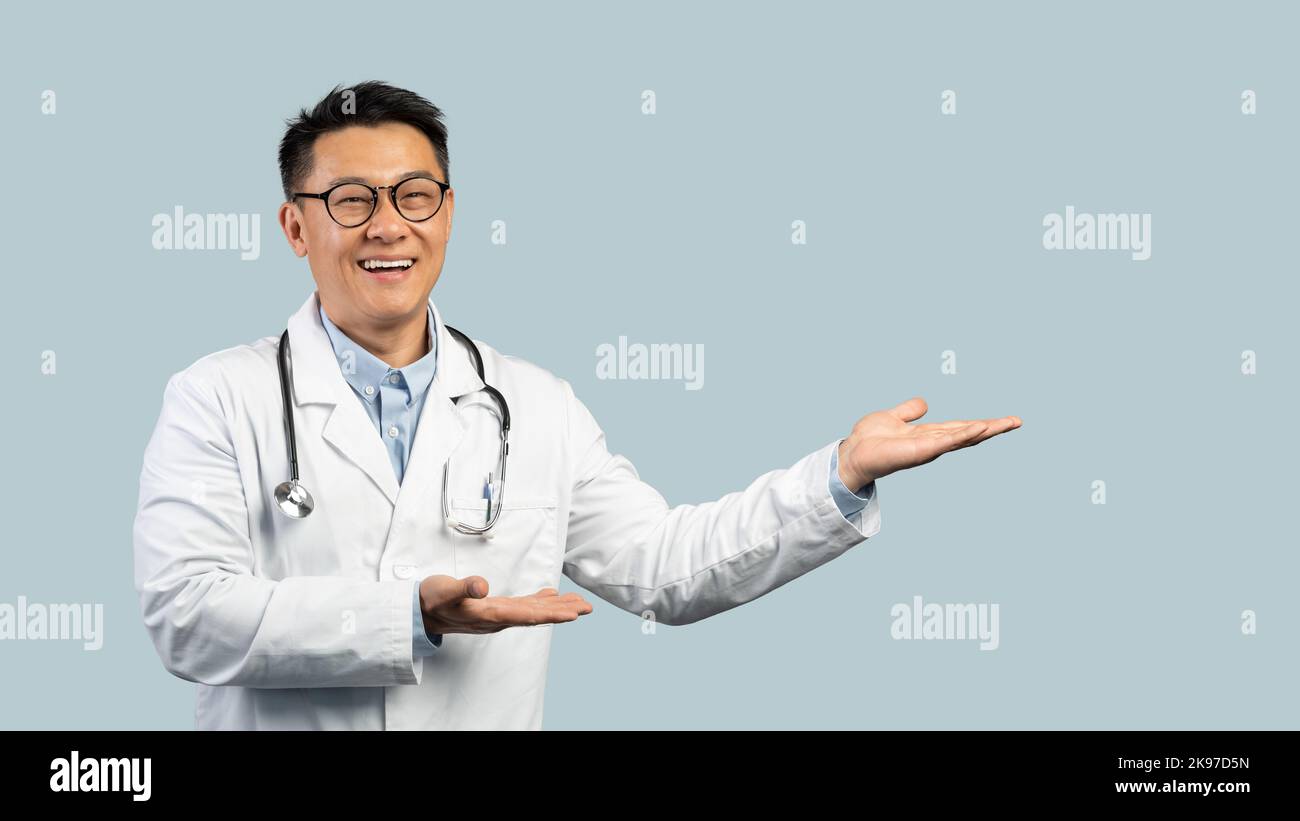 Joyeux homme chinois mature médecin en manteau blanc, les lunettes pointent les mains vers l'espace vide Banque D'Images