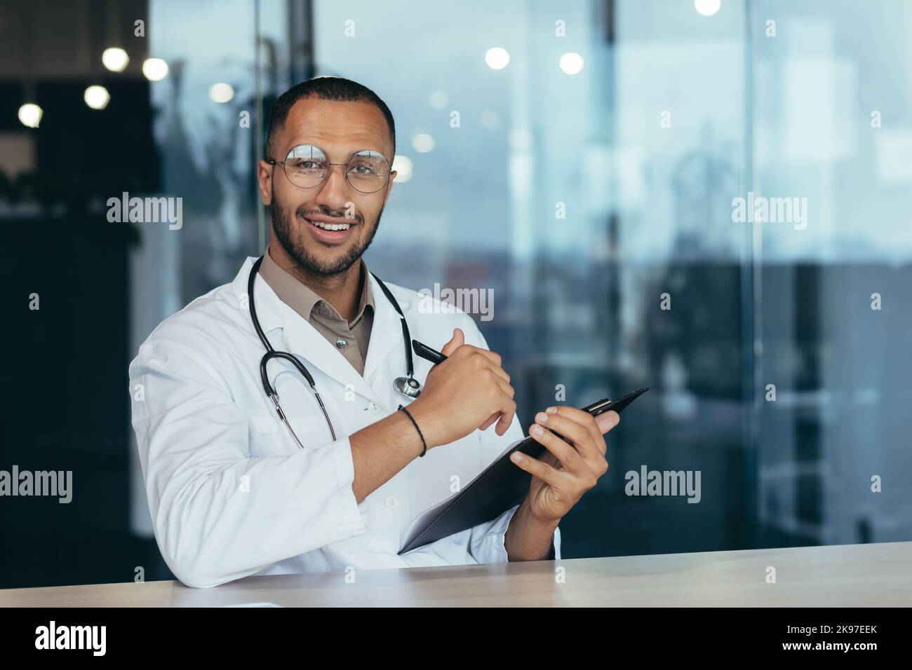 Portrait d'un jeune beau médecin latino-américain. Assis dans le bureau, tenant un stylo avec des documents dans ses mains, regardant la caméra, souriant Banque D'Images