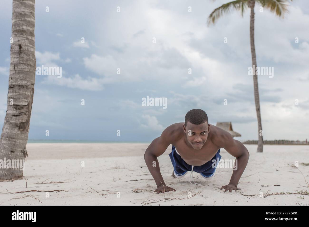 Athlète américain africain musclé dans les vêtements de sport en se poussant vers le haut tout en s'entraînement sur la plage de sable sous ciel nuageux et en regardant la caméra Banque D'Images